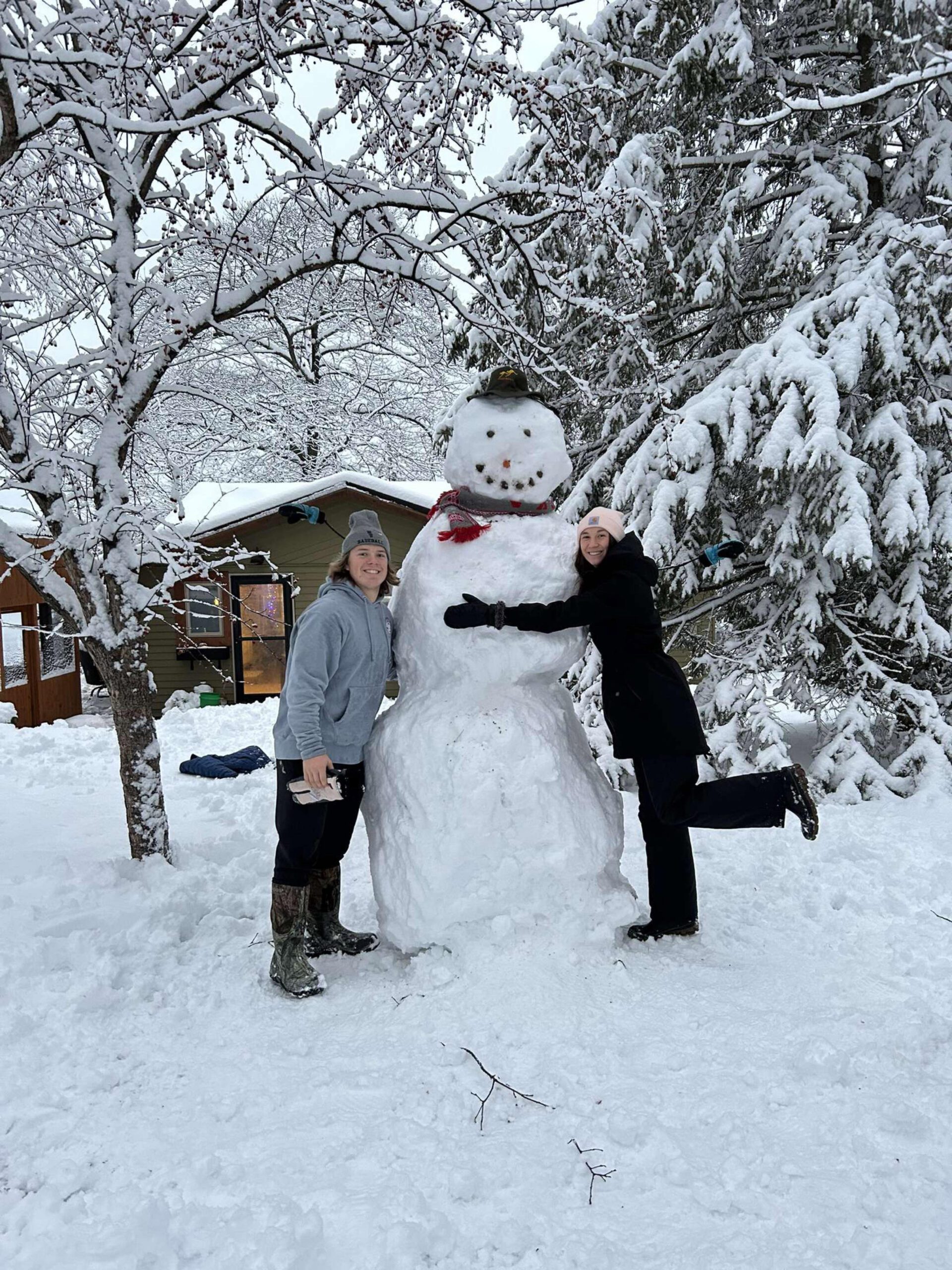 Tall snowman with black hat being hugged by girl and a boy stands on the left