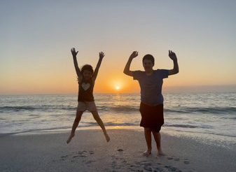 A boy and a girl jump on the beach with the Gulf and sunset behind.
