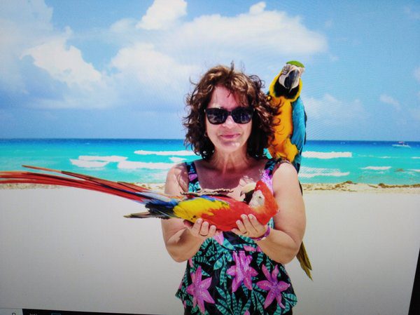 Woman in a floral swimsuit hold a red parrot in her hand, another blue bird on her shoulder.