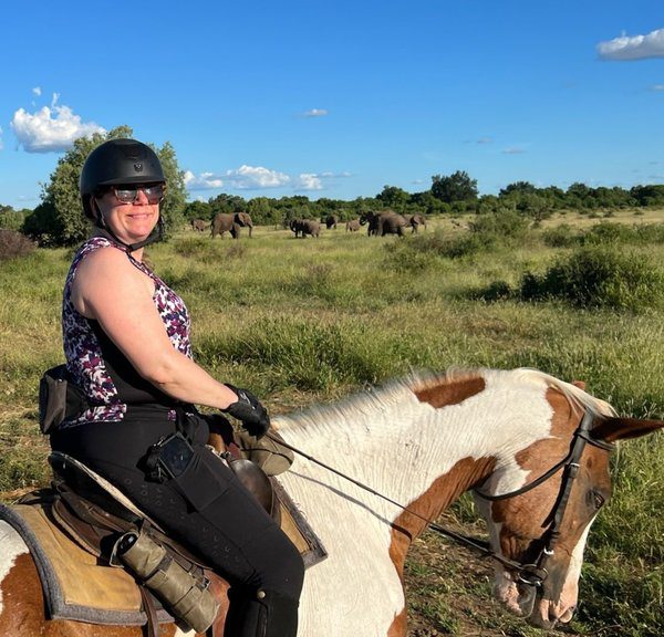 Woman on a horse, smiling at the camera with a herd of elephants way in the back.