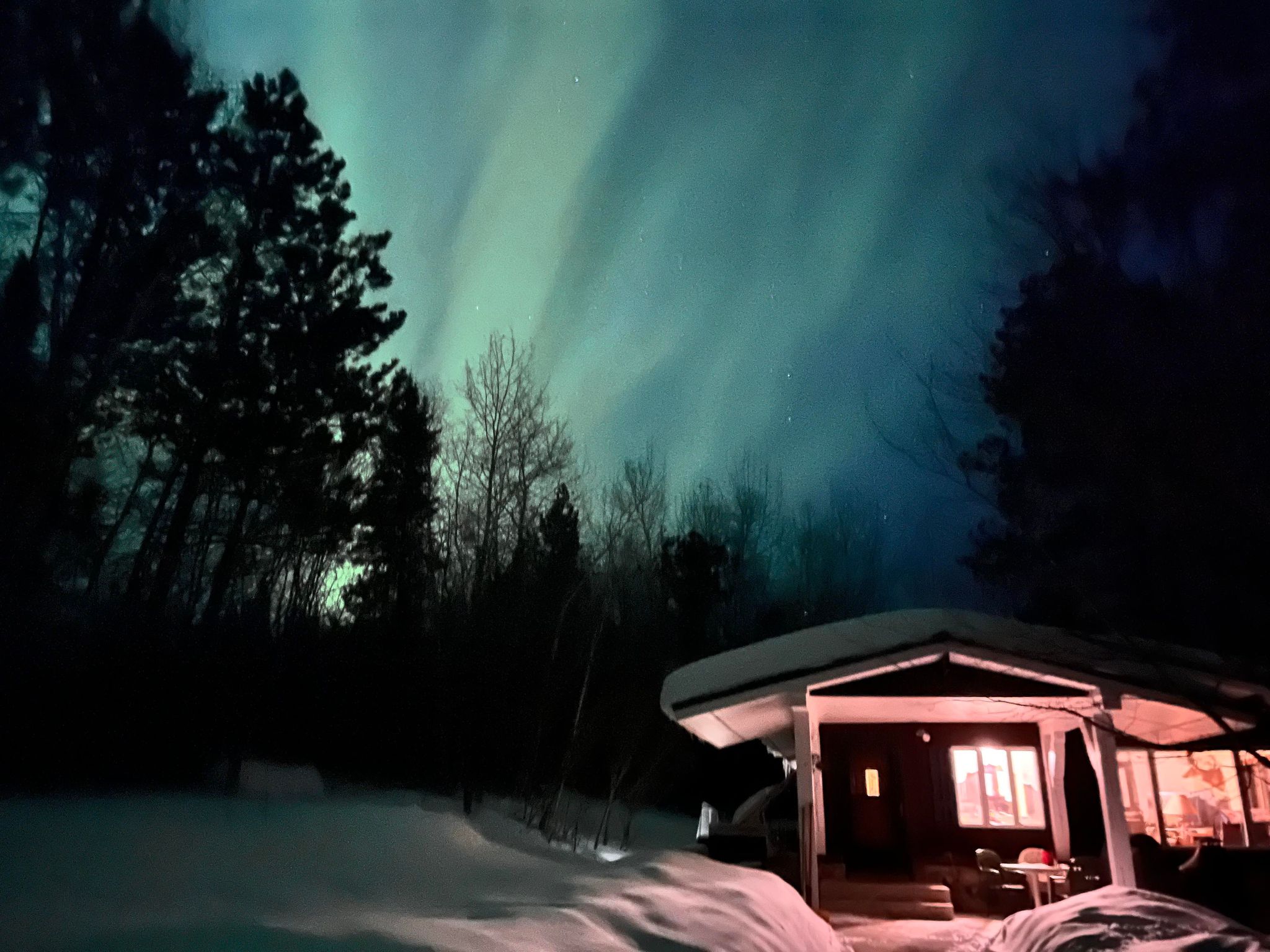 A lighted house with a porch, covered in snow with the Northern Lights streaking blue and green.